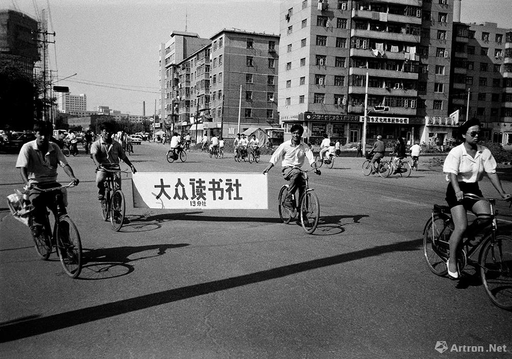 谢军作品：1994.8 沈阳 青年大街 两青年人骑着车拿着要开张的“大众读书社19分社”的牌匾经过城市最宽的马路。